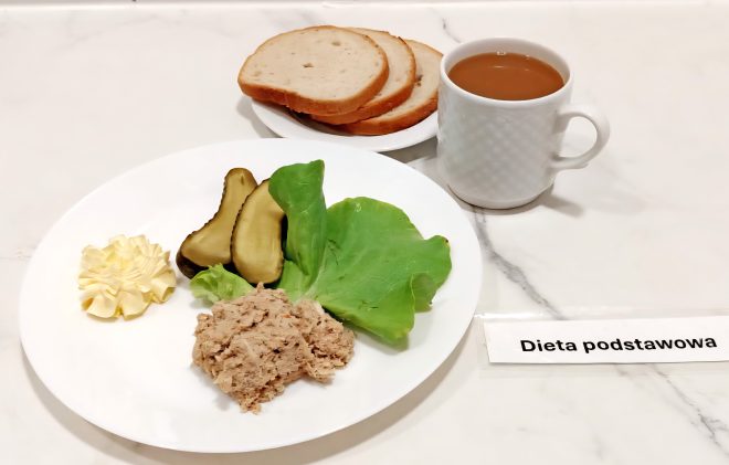 Zestaw śniadaniowy w diecie podstawowej podany na białym talerzu. W skład posiłku wchodzą: pasta mięsna, dwa plasterki ogórka konserwowego, liść sałaty oraz porcja masła. W tle widoczny talerzyk z trzema kromkami białego pieczywa oraz biały kubek z kawą z mlekiem. Kompozycja znajduje się na jasnym marmurowym blacie, obok etykieta z napisem „Dieta podstawowa”