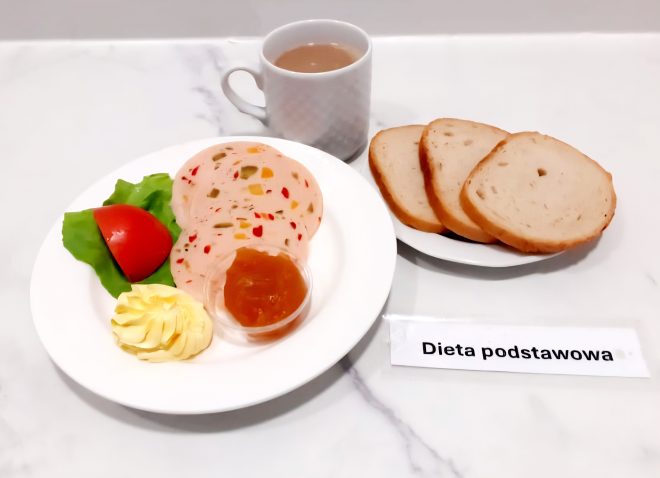 Talerz z plasterkami galarety drobiowej z warzywami, liściem sałaty, pomidorem, masłem i porcją dżemu. Obok trzy kromki pieczywa pszennego oraz filiżanka kawy z mlekiem. Dieta podstawowa