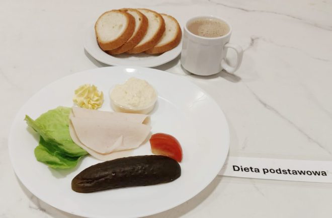 Śniadanie złożone z kilku kromek białego pieczywa, dwóch plasterków wędliny drobiowej, liścia sałaty, kawałka pomidora, dużego ogórka kiszonego, porcji masła, pasty jajecznej i filiżanki kawy z mlekiem. Na talerzu etykieta: „Dieta podstawowa”