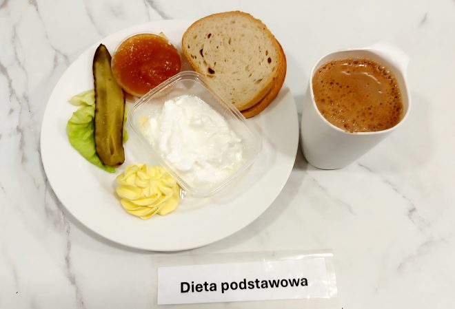 Zestaw śniadaniowy w ramach diety podstawowej. Na białym talerzu znajdują się trzy kromki pieczywa pszennego, porcja białego twarożku w plastikowym pojemniczku, dekoracyjnie wyciśnięta porcja masła, plaster kiszonego ogórka ułożony na liściu sałaty oraz porcja marmolady z jasnych owoców (najprawdopodobniej morelowej). Napój do śniadania – kakao lub kawa zbożowa z mlekiem – został podany w białym ceramicznym kubku. Posiłek ułożono na marmurowym blacie z etykietą „Dieta podstawowa”