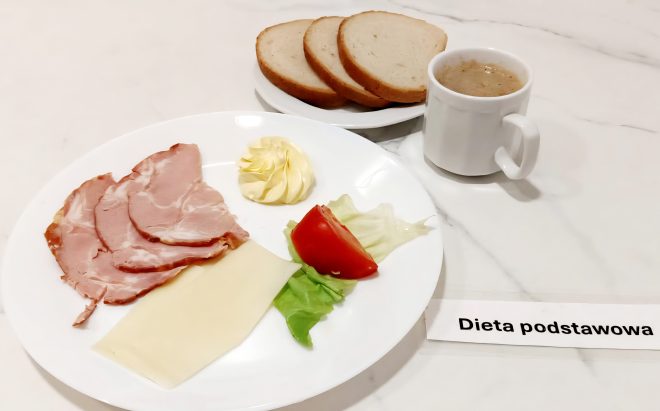 Biały talerz z trzema plasterkami szynki, plastrem żółtego sera, dekoracyjną porcją masła, liściem sałaty i kawałkiem pomidora. W tle talerzyk z bułką kajzerką i dwoma kromkami chleba, dwa kubki – jeden z kawą zbożową, drugi z mlekiem z płatkami. Na stoliku etykieta: "Dieta podstawowa"