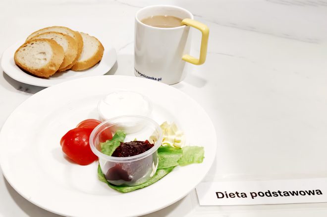 Śniadanie serwowane na białym marmurowym blacie. Na pierwszym talerzu – trzy kromki pszennego pieczywa. Na drugim, większym talerzu – porcja masła, dwa plasterki pomidora, kilka liści sałaty, dwie małe przezroczyste miseczki: jedna zawierająca biały nabiał (prawdopodobnie twarożek lub jogurt naturalny), druga – porcję dżemu wiśniowego lub śliwkowego. Do posiłku dołączony biały kubek z herbatą lub kawą z mlekiem. Całość oznaczona karteczką z napisem „Dieta podstawowa”