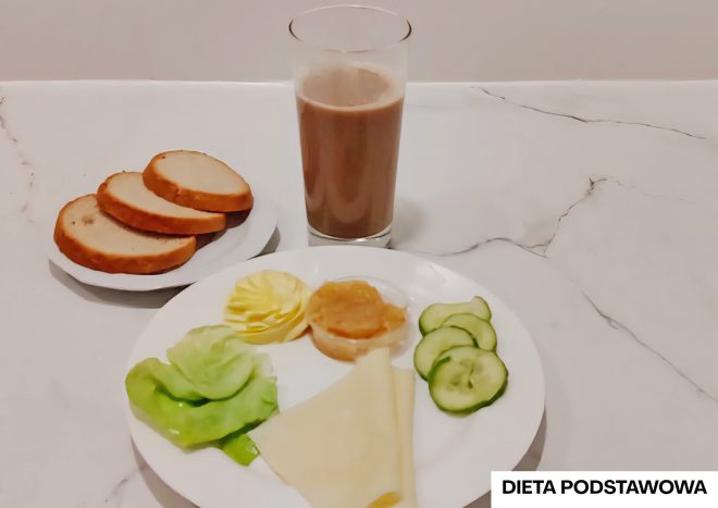 Talerz z trzema plastrami ogórka, liśćmi sałaty, plastrem żółtego sera, porcją masła i małym kubeczkiem dżemu jabłkowego. Obok trzy kromki białego pieczywa i szklanka kakao. W prawym dolnym rogu etykieta z napisem „Dieta podstawowa”
