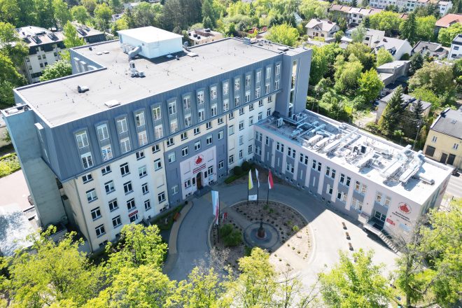 Widok z lotu ptaka na nowoczesny budynek Szpitala Zakonu Bonifratrów św. Jana Grandego w Krakowie, otoczony zielenią i niską zabudową mieszkalną. Na dziedzińcu przed głównym wejściem powiewają cztery flagi: papieska, szpitalna, polska i bonifraterska. Na ścianie frontowej widoczne logo Zakonu Bonifratrów oraz napis 