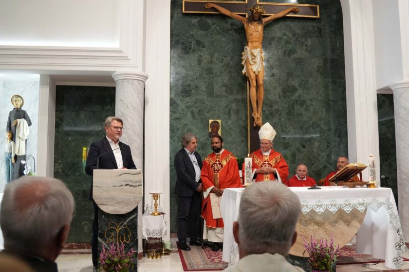 Uroczysta Msza Święta w kaplicy św. Ryszarda Pampuri. Mężczyzna w garniturze przemawia z ambony, a za nim stoją brat Saji, biskup Michał Janocha oraz inni duchowni. W tle duży krucyfiks