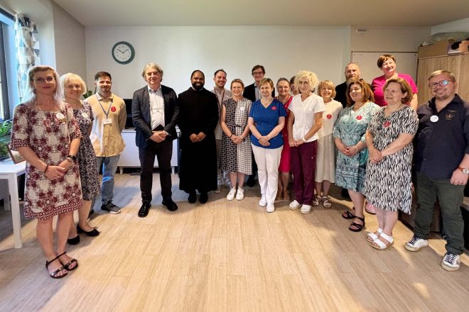 Grupowe zdjęcie pracowników Bonifraterskiego Centrum Medycznego w Warszawie z Wizytatorem Generalnym bratem Saji Mullankuzhy OH. Uczestnicy stoją razem w pomieszczeniu administracyjnym, uśmiechnięci, z serduszkowymi przypinkami