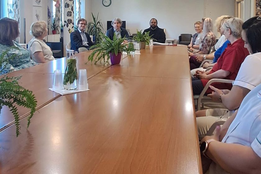 Spotkanie pracowników z bratem Saji Mullankuzhy OH przy dużym stole konferencyjnym. Wnętrze ozdobione zielonymi roślinami i ziołową wodą na stole. W tle zegar i dekoracje ścienne