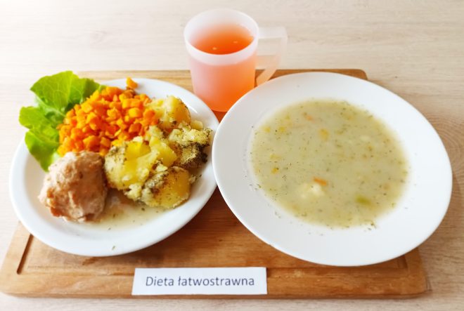 Taca z posiłkiem oznaczonym jako „Dieta łatwostrawna”. Zawiera dwa talerze i przezroczysty kubek z jasnoczerwonym napojem. Na dużym talerzu zupa jarzynowa z koperkiem. Na mniejszym talerzu: gotowany ziemniak posypany ziołami, gotowana marchewka w kostkę, pulpet drobiowy oraz liść sałaty