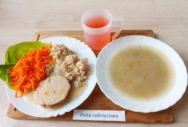 Talerz obiadowy z daniem w diecie cukrzycowej – plaster pieczeni w sosie, porcja kaszy oraz surówka z tartej marchewki na liściu sałaty. Obok znajduje się talerz z zupą jarzynową na bazie bulionu i kubek z różowym napojem. Całość na tacy z podpisem „Dieta cukrzycowa”