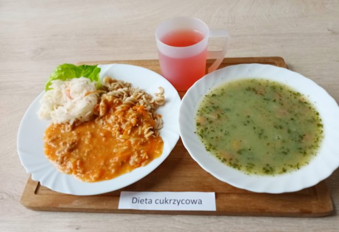Zestaw obiadowy na drewnianej tacy. Na jednej połowie tacy znajduje się talerz z makaronem w sosie pomidorowym z warzywami, obok surówka z kapusty oraz liść sałaty. Na drugiej połowie tacy stoi miska z zieloną zupą jarzynową z ziołami. Do picia – różowy napój w plastikowym kubku z uchem. Pod spodem widoczny napis: „Dieta cukrzycowa”