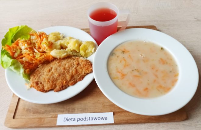Talerz z obiadem w diecie podstawowej: zupa z tartą marchewką i ziemniakami, drugie danie z kotletem schabowym, ziemniakami oraz surówką z marchewki i kapusty, podanymi na liściu sałaty. Obok kubek z czerwonym kompotem. Całość serwowana na drewnianej tacy z etykietą "Dieta podstawowa"