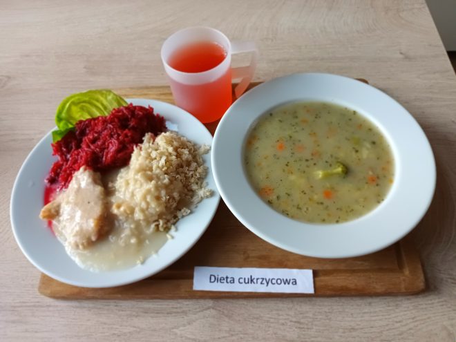 Taca z obiadem oznaczonym jako „Dieta cukrzycowa”. Na niej znajduje się miska z zupą jarzynową (z widocznymi kawałkami marchewki, brokuła i koperku) oraz talerz z kaszą, duszonym mięsem w jasnym sosie, buraczkami na sałacie i różowym napojem w plastikowym kubku