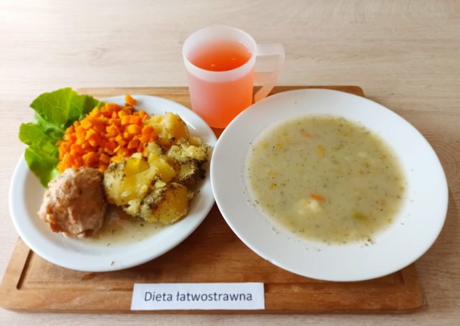 Na drewnianej tacy znajduje się zestaw obiadowy opisany jako „Dieta łatwostrawna”. W skład posiłku wchodzą: talerz z jasną, klarowną zupą jarzynową z widocznymi drobnymi kawałkami warzyw oraz talerz z gotowanym ziemniakiem z koperkiem, gotowaną marchewką w kostkę, pulpą mięsną i liściem sałaty jako dekoracją. Do posiłku podano także kubek różowego napoju owocowego