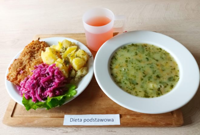Zestaw obiadowy w diecie podstawowej. Na tacy umieszczono talerz z zupą jarzynową z natką pietruszki, a obok drugi talerz z panierowanym kotletem schabowym, gotowanymi ziemniakami z koperkiem oraz surówką z czerwonej kapusty na liściu sałaty. Do obiadu podano czerwony napój w przezroczystym kubku. Całość ustawiona na tacy z etykietą „Dieta podstawowa”