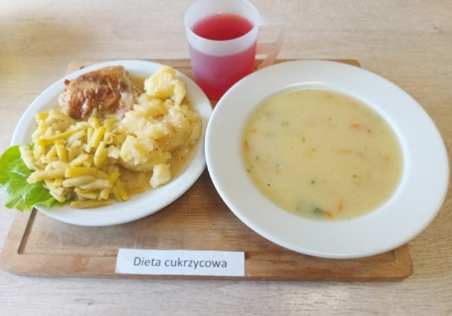 Obiad w ramach diety cukrzycowej podany na tacy. Na talerzu widoczna porcja gotowanych ziemniaków z sosem, duszone udko z kurczaka oraz gotowana fasolka szparagowa na liściu sałaty. Obok talerz z zupą jarzynową oraz kubek z czerwonym kompotem. Posiłek oznaczony kartką z napisem „Dieta cukrzycowa”