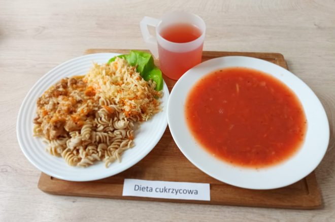Zestaw obiadowy w diecie cukrzycowej. Na tacy znajdują się dwa talerze: jeden z zupą pomidorową z makaronem oraz drugi z porcją mięsa w sosie, gotowanym makaronem świderki razowego typu i surówką z kiszonej kapusty i marchwi. Obok plastikowy kubek z czerwonym napojem owocowym