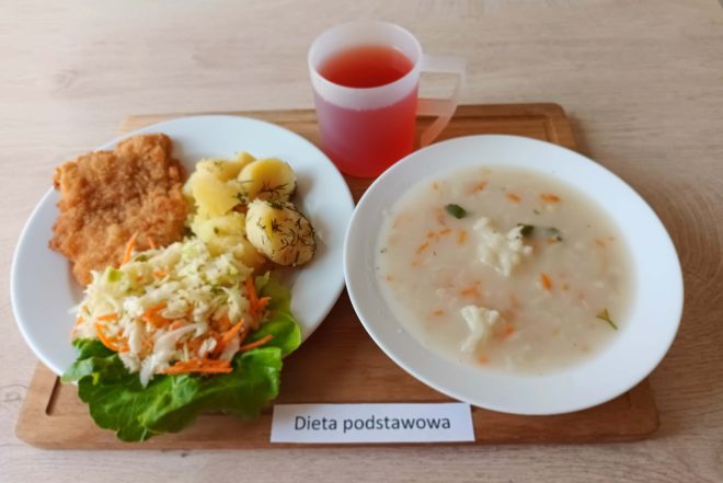 Na tacy znajduje się obiad z diety podstawowej. Po lewej stronie talerza leży panierowany kotlet schabowy, obok porcja ziemniaków posypanych świeżym koperkiem oraz surówka z kapusty pekińskiej z marchwią, ułożona na liściu sałaty. Po prawej stronie miska z zupą jarzynową na białym wywarze – widać kawałki kalafiora, marchewki i zieloną pietruszkę. Obok plastikowy kubek z czerwonym kompotem. Wszystko podane na drewnianej tacy, z podpisem „Dieta podstawowa”