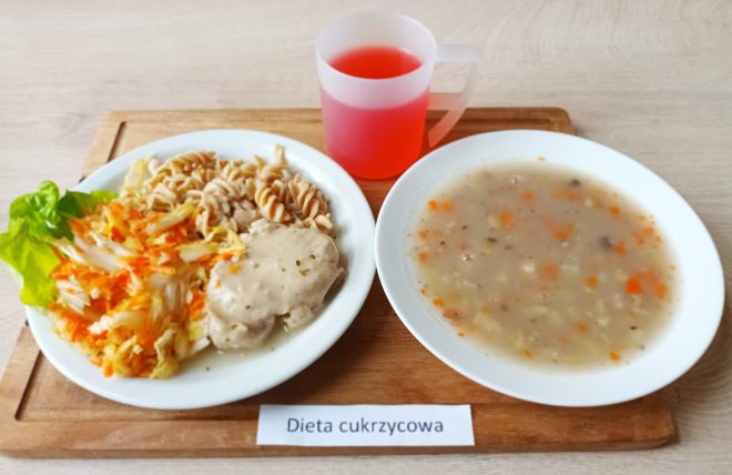 Taca z posiłkiem oznaczonym jako „Dieta cukrzycowa”. Na białym talerzu obiadowym znajduje się porcja gotowanego makaronu razowego, kawałek mięsa w jasnym sosie oraz surówka z kapusty pekińskiej i marchwi, podana na liściu sałaty. Obok stoi talerz z zupą jarzynową z widocznymi kawałkami marchewki, ziemniaka i kaszy. W przezroczystym kubku z matowego plastiku znajduje się czerwony napój owocowy. Całość umieszczona na drewnianej tacy. Zdjęcie wykonane na jasnym blacie stołu