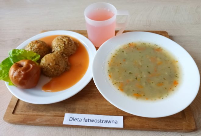 Zupa jarzynowa w białym talerzu oraz pulpety z kaszy w sosie pomidorowym z dodatkiem pieczonego jabłka i listka sałaty. Do picia różowy napój w przezroczystym plastikowym kubku. Posiłek w ramach diety łatwostrawnej.