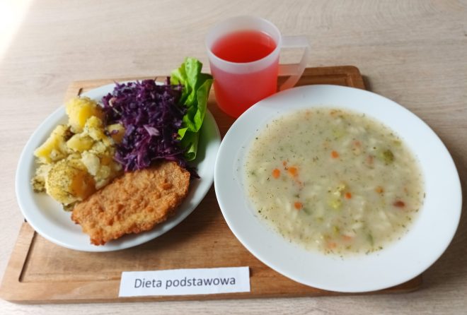 Obiad w diecie podstawowej na białych naczyniach – zupa jarzynowa z ryżem i koperkiem oraz drugie danie: panierowany filet (najprawdopodobniej drobiowy), gotowane ziemniaki z koperkiem, surówka z czerwonej kapusty i liść sałaty. Do picia czerwony napój owocowy w plastikowym kubku. Na desce napis: „Dieta podstawowa”