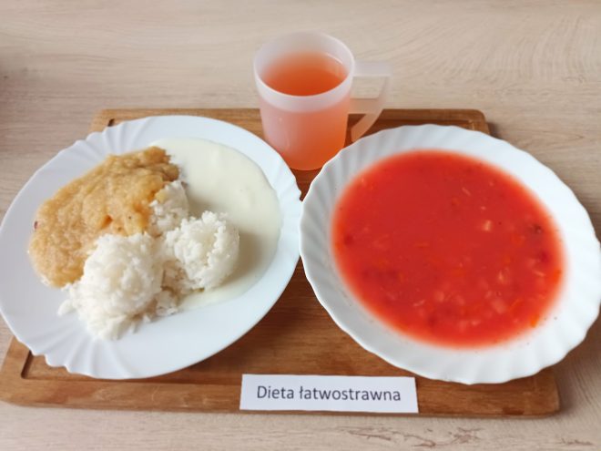 Na tacy drewnianej ustawiono trzy elementy: talerz z czerwonym barszczem jarzynowym, talerz z gotowanym ryżem polanym białym sosem i musem jabłkowym, oraz kubek z jasnoczerwonym napojem owocowym. Pod tacą znajduje się podpis: „Dieta łatwostrawna”