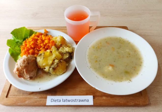 Dwukomorowe pudełko z ziemniakami posypanymi koperkiem oraz mielonym kotletem. W osobnych pojemnikach – starta surówka z marchewki oraz zupa pomidorowa z ryżem. Do posiłku szklanka napoju (prawdopodobnie herbata). Całość ustawiona na desce z etykietą „Dieta podstawowa”