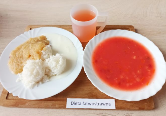 Taca z obiadem dla osoby na diecie łatwostrawnej. Na jednym talerzu znajduje się gotowany ryż z musem jabłkowym i jasnym sosem. Obok talerz z klarowną zupą jarzynową o czerwonym kolorze (prawdopodobnie barszcz lub pomidorowa z drobnymi warzywami). Do posiłku podano także kubek z czerwonym napojem. Etykieta na tacy brzmi: „Dieta łatwostrawna”