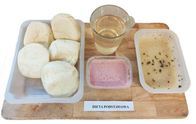 Taca z posiłkiem oznaczonym jako „Dieta podstawowa”. W skład posiłku wchodzi pięć jasnych bułek pszennych w plastikowym pojemniku, różowy jogurt lub mus owocowy w małym przezroczystym pojemniczku, szklanka jasnego napoju (np. herbaty lub naparu), oraz kremowa zupa lub kaszka posypana przyprawami, podana w prostokątnym pojemniku
