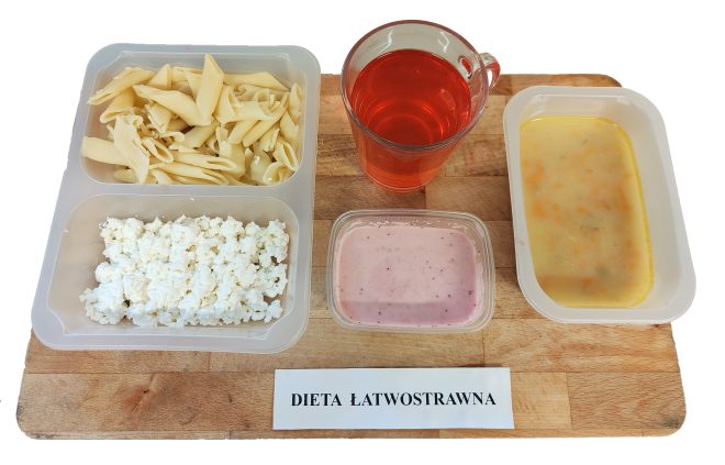 Komplet obiadowy dla diety łatwostrawnej: makaron penne bez dodatków, twarożek, różowy jogurt owocowy, klarowna zupa warzywna z widoczną marchewką oraz czerwony napój owocowy w szklanym kubku. Wszystko ułożone na desce z podpisem „Dieta łatwostrawna”