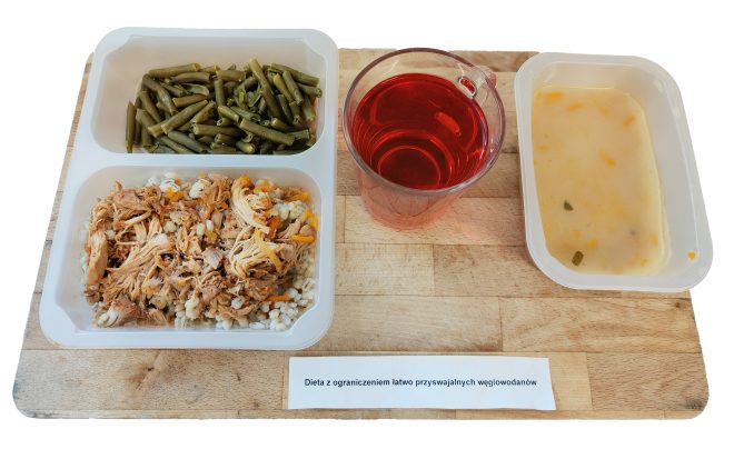 Taca obiadowa z daniem zgodnym z dietą niskowęglowodanową. W pojemniku: duszone mięso z warzywami podane na jęczmieniu perłowym, gotowana fasolka szparagowa. Obok kubek z czerwonym naparem oraz pojemnik z jasną zupą jarzynową