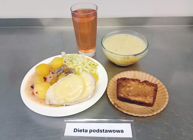 Taca z obiadem w ramach „Diety podstawowej”. Na niej: talerz z gotowanymi ziemniakami, białą kapustą zasmażaną lub surówką oraz mięsem z plastrem roztopionego sera i sosem z pieczarkami. Obok głębokiego talerza znajduje się zupa jarzynowa lub ziemniaczana z koperkiem, a także kromka ciasta na papierowej podkładce i wysoki szklany kubek z herbatą lub kompotem