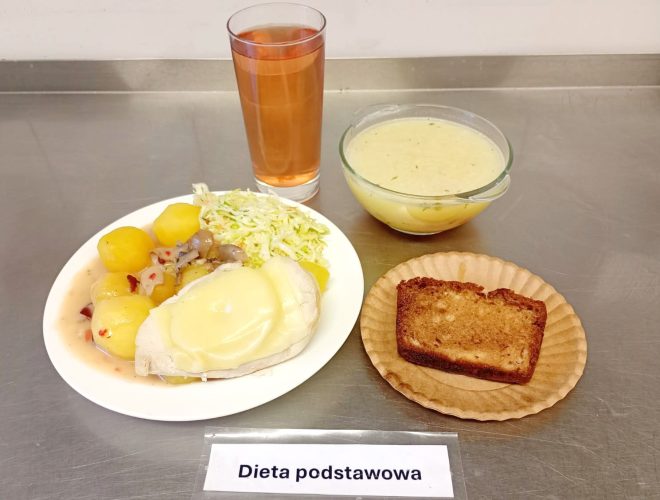 Taca z obiadem w ramach diety podstawowej. Na talerzu gotowane ziemniaki, gotowany filet z kurczaka zapieczony z żółtym serem, sos z pieczarkami oraz surówka z białej kapusty. Obok talerza miseczka z lekką zupą ziemniaczaną lub koperkową, szklanka czerwonego kompotu oraz kawałek ciasta domowego w papierowej foremce. Całość na metalowym blacie z etykietą „Dieta podstawowa”