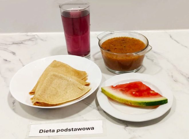 Na marmurowym blacie ustawiono komplet posiłku w ramach diety podstawowej. Na pierwszym planie znajduje się biały talerz z trzema złożonymi na trójkąty naleśnikami. Obok stoi miseczka z przezroczystego szkła, wypełniona zupą warzywną z widocznymi kawałkami warzyw i przypraw. Dodatkiem jest soczysty trójkątny kawałek arbuza oraz szklanka ciemnoróżowego kompotu. Całość wygląda lekko, letnio i apetycznie