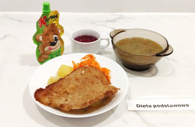 Talerz obiadowy z panierowanym kotletem schabowym, gotowanymi ziemniakami i surówką z marchewki. Obok znajduje się filiżanka z kompotem, saszetka owocowego musu w tubce oraz miseczka z zupą jarzynową. Całość oznaczona karteczką z napisem "Dieta podstawowa"