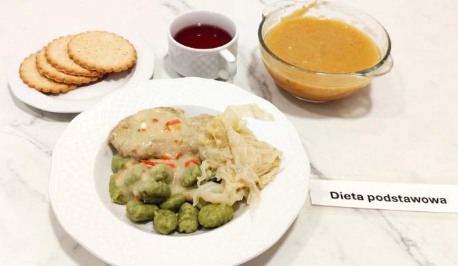Talerz z obiadem w ramach diety podstawowej, na którym znajdują się zielone kopytka ze szpinakiem polane jasnym sosem, kotlet mielony w sosie z papryką oraz duszona biała kapusta. Obok znajdują się: filiżanka z czerwonym kompotem, miseczka z zupą jarzynową i talerzyk z sześcioma kruchymi herbatnikami. Na stole widoczna kartka z napisem "Dieta podstawowa"