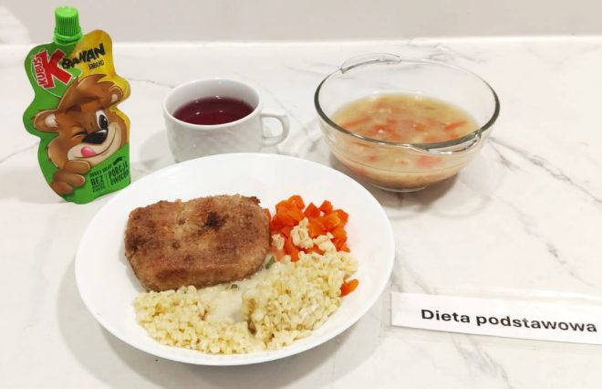 Taca z obiadem oznaczonym jako „Dieta podstawowa”. Na talerzu panierowany kotlet, kasza jęczmienna z jasnym sosem, gotowana marchewka i kalafior. Obok szklana miseczka z zupą warzywną z ziemniakami i marchewką, kubek z czerwonym kompotem oraz mus owocowy Kubuś o smaku banan-jabłko w tubce