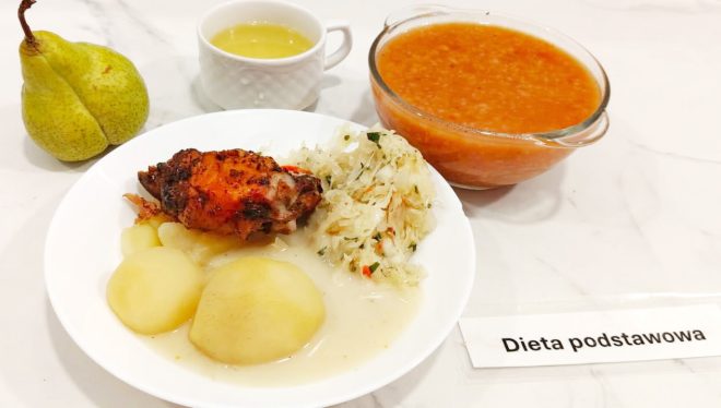 Pełnowartościowy obiad z diety podstawowej: porcja pieczonego kurczaka z przypieczoną skórką, gotowane ziemniaki z sosem, surówka z kiszonej kapusty z marchewką i natką pietruszki. Obok na stole gruszka, filiżanka z jasnym kompotem oraz miska z zupą pomidorową z ryżem. Posiłek podany na białym talerzu, na jasnym blacie, obok karteczka z napisem „Dieta podstawowa”