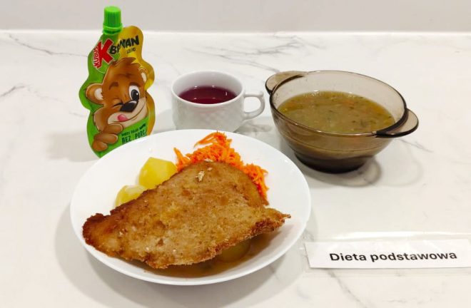 Obiad w diecie podstawowej, podany na białym tle. Na talerzu głównym znajdują się gotowane ziemniaki, surówka z tartej marchwi oraz duży kotlet schabowy w panierce. W tle kubek z czerwonym naparem (kompot lub herbata) oraz szklana miska z zupą jarzynową na wywarze ziołowym – widać kawałki warzyw i natki pietruszki. Po lewej stronie opakowanie musu owocowego Kubuś. Obok karteczka z napisem „Dieta podstawowa”
