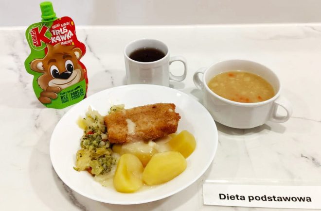 Obiad w diecie podstawowej, sfotografowany na jasnym blacie. Na talerzu ziemniaki, porcja warzyw duszonych (z kapustą, marchewką, brokułami) oraz panierowany filet rybny z sosem. Obok filiżanka z ciemnym naparem i miseczka z zupą warzywną z pływającymi kawałkami marchwi i ziemniaków. Z lewej strony widoczny mus warzywno-owocowy Kubuś. W prawym dolnym rogu kartka z napisem „Dieta podstawowa”