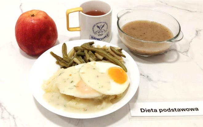 Posiłek z diety podstawowej serwowany na białym talerzu. Na talerzu znajdują się dwa jajka sadzone na puree ziemniaczanym, polane jasnym sosem koperkowym. Obok porcja gotowanej fasolki szparagowej. Z boku ustawiono czerwone jabłko oraz kubek z herbatą w białym naczyniu z żółtym uchem i logotypem "Kryniczanka". Obok szklana miseczka z zupą jarzynową. Wszystko ustawione na jasnym, marmurowym blacie