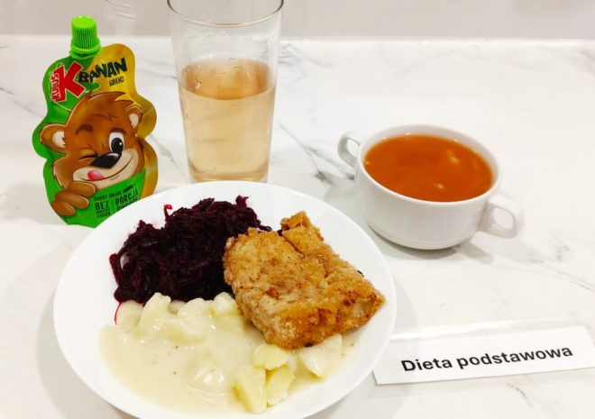 Obiad w diecie podstawowej – panierowany filet rybny z gotowanymi ziemniakami polanymi sosem, podany z buraczkami ćwikłowymi. Do zestawu dołączona zupa pomidorowa w białej filiżance, szklanka napoju oraz mus owocowy w saszetce „Kubuś Banan Jabłko”. Posiłek serwowany na marmurowym blacie z etykietą „Dieta podstawowa”