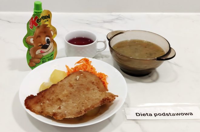 a zdjęciu przedstawiony jest zestaw obiadowy przeznaczony dla pacjentów stosujących dietę podstawową. Składa się z: miski zupy – prawdopodobnie jarzynowej lub ogórkowej z ziemniakami i zieleniną, serwowanej w przezroczystym naczyniu, dania głównego na białym talerzu: panierowanego kotleta schabowego lub drobiowego, gotowanych ziemniaków oraz surówki z marchwi, saszetki musu owocowego „Kubuś” o smaku bananowym, filiżanki z herbatą owocową. Całość ułożona na białym blacie z oznaczeniem: „Dieta podstawowa”