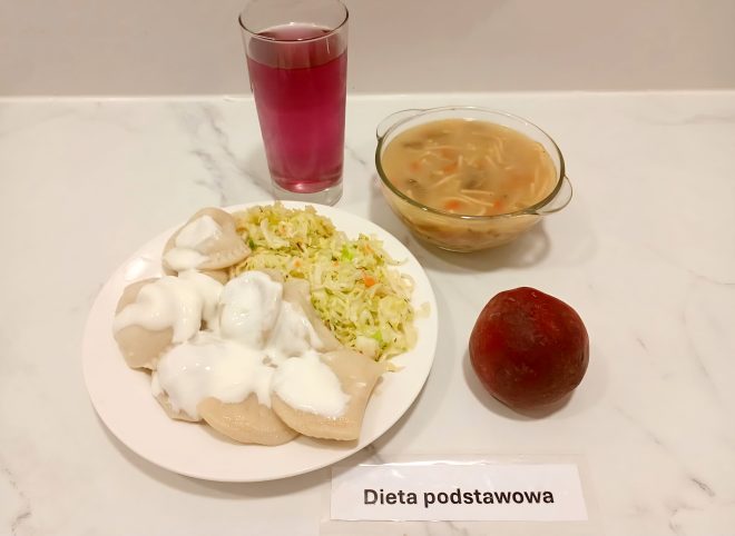 Taca z obiadem oznaczonym jako „Dieta podstawowa”. Na talerzu pierogi z białym serem polane śmietaną oraz surówka z kiszonej kapusty z marchewką. Obok zupa jarzynowa z makaronem w szklanej miseczce, różowy napój w wysokiej szklance i czerwony owoc – najprawdopodobniej jabłko