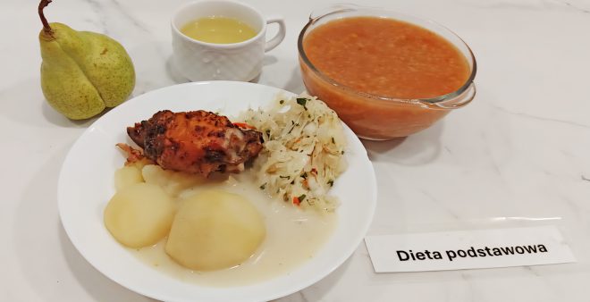 Talerz obiadowy w diecie podstawowej: pieczone udko z kurczaka, gotowane ziemniaki polane lekkim sosem oraz surówka z białej kapusty z marchewką i natką pietruszki. Obok talerza gruszka, filiżanka z jasnym kompotem oraz miska z pomarańczową zupą jarzynową. Obok posiłku karteczka z napisem „Dieta podstawowa”