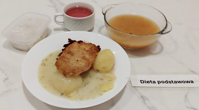 Talerz z dwoma ziemniakami w sosie koperkowym, panierowanym kotletem schabowym oraz dodatkiem buraczków. W tle kompot wiśniowy, miseczka z lekką zupą oraz deser w przezroczystym pojemniku. Całość oznaczona etykietą „Dieta podstawowa”