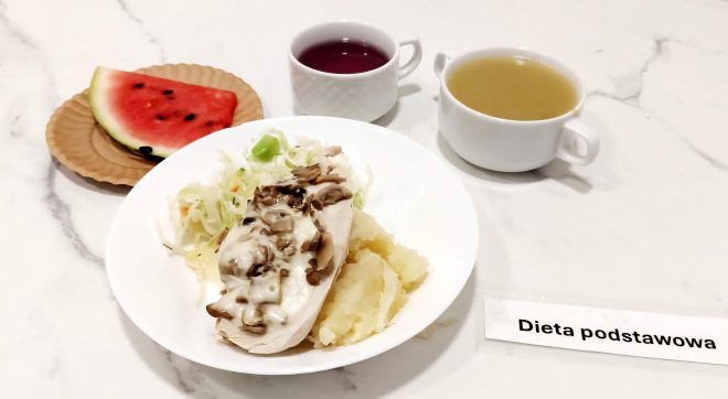 Zestaw obiadowy składający się z gotowanych ziemniaków, duszonej piersi kurczaka z pieczarkami i serem oraz surówki z kapusty i warzyw. W tle dwa napoje: filiżanka z kompotem wiśniowym i kubek z lekkim rosołem. Całość uzupełnia porcja świeżego arbuza na papierowym talerzyku. Kompozycja estetycznie ułożona na jasnym blacie z widoczną etykietą „Dieta podstawowa”