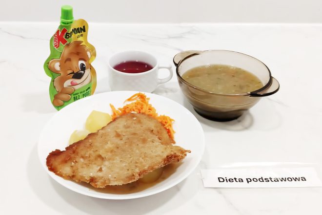 Obiad w diecie podstawowej: na talerzu ziemniaki, starta marchewka i duży panierowany kotlet schabowy. Obok przezroczysta miseczka z zupą oraz kubek z czerwonym napojem owocowym. Z boku opakowanie owocowego musu typu „Kubuś” z wizerunkiem misia. Na stole kartka z napisem „Dieta podstawowa”