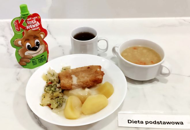 Obiad w diecie podstawowej, podany na marmurowym blacie. Na białym talerzu znajdują się gotowane ziemniaki, warzywa duszone z kapustą i brokułami oraz panierowany filet rybny z jasnym sosem. Obok filiżanka z ciemnym naparem (najpewniej herbata) i miseczka z zupą jarzynową – w bulionie widoczne są kawałki ziemniaków i marchwi. Po lewej stronie stoi mus warzywno-owocowy Kubuś. Przed talerzem kartka z podpisem „Dieta podstawowa”