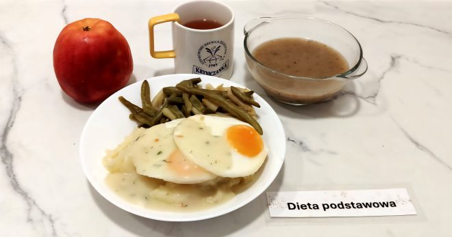 Posiłek obiadowy w diecie podstawowej. Na białym talerzu znajduje się porcja puree ziemniaczanego, jajka sadzonego z sosem koperkowym oraz gotowana fasolka szparagowa. Obok ustawiono czerwone jabłko, przezroczysty kubek z herbatą z logo „Kryniczanka” oraz szklana miseczka z jasną zupą jarzynową. Zdjęcie wykonane na białym marmurowym blacie, z widoczną etykietą „Dieta podstawowa”