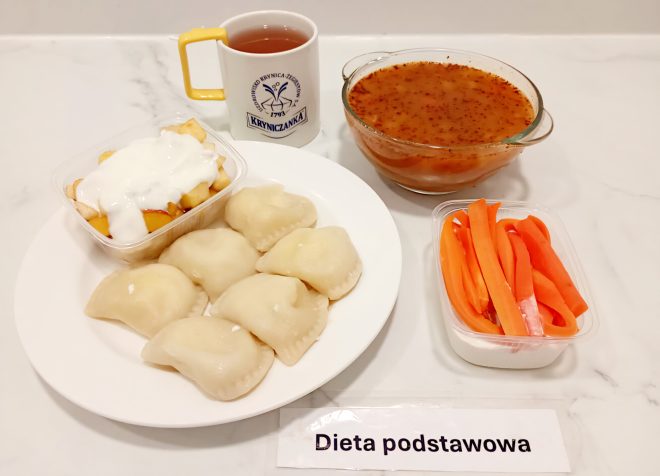 Taca z obiadem szpitalnym oznaczonym jako „Dieta podstawowa”. Na talerzu znajduje się porcja pierogów z serem lub ziemniakami. Obok w plastikowym pojemniku zapiekane jabłka z jogurtem naturalnym lub śmietaną. W osobnej miseczce czerwony barszcz z ziemniakami i przyprawami. W oddzielnym pojemniku paski surowej marchewki. Do posiłku podano także kubek herbaty w białym kubku z logo „Kryniczanka”